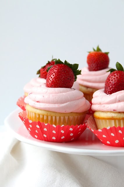 Strawberry Cupcakes with Fresh Strawberry Buttercream are the perfect summer dessert, packed with fresh strawberries. You'll love this easy cupcakes recipe! #cupcakes #recipe #strawberries #desserts cake plate holding homemade strawberry cupcakes topped with strawberry buttercream and whole fresh strawberries