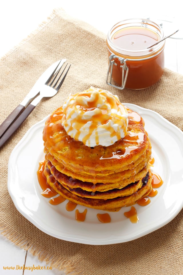 A stack of pumpkin spice pancakes topped with whipped cream and caramel drizzle on a white plate