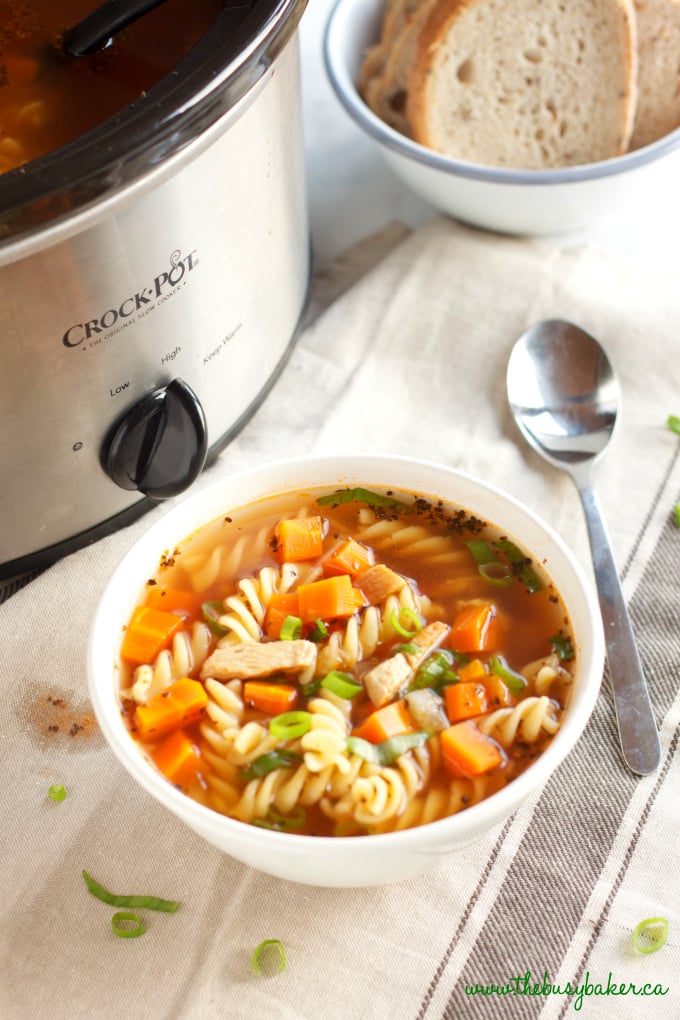 pasta, carrots, chicken in a broth in a white bowl with a crock pot in the background