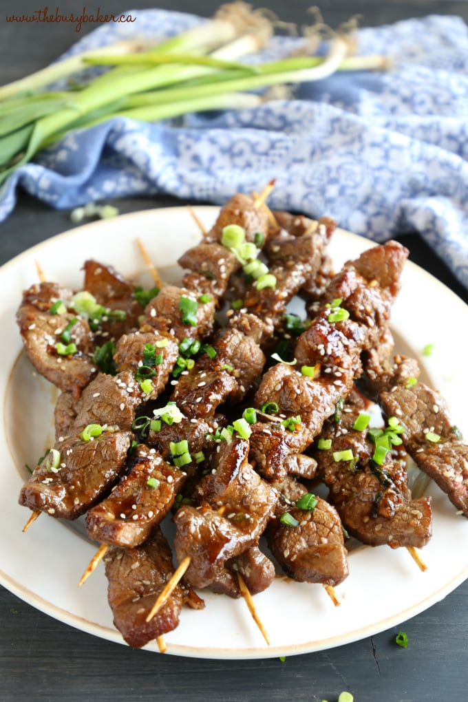 Best Ever Korean Barbecue Beef Skewers on white plate with green onions and blue cloth and sesame seeds