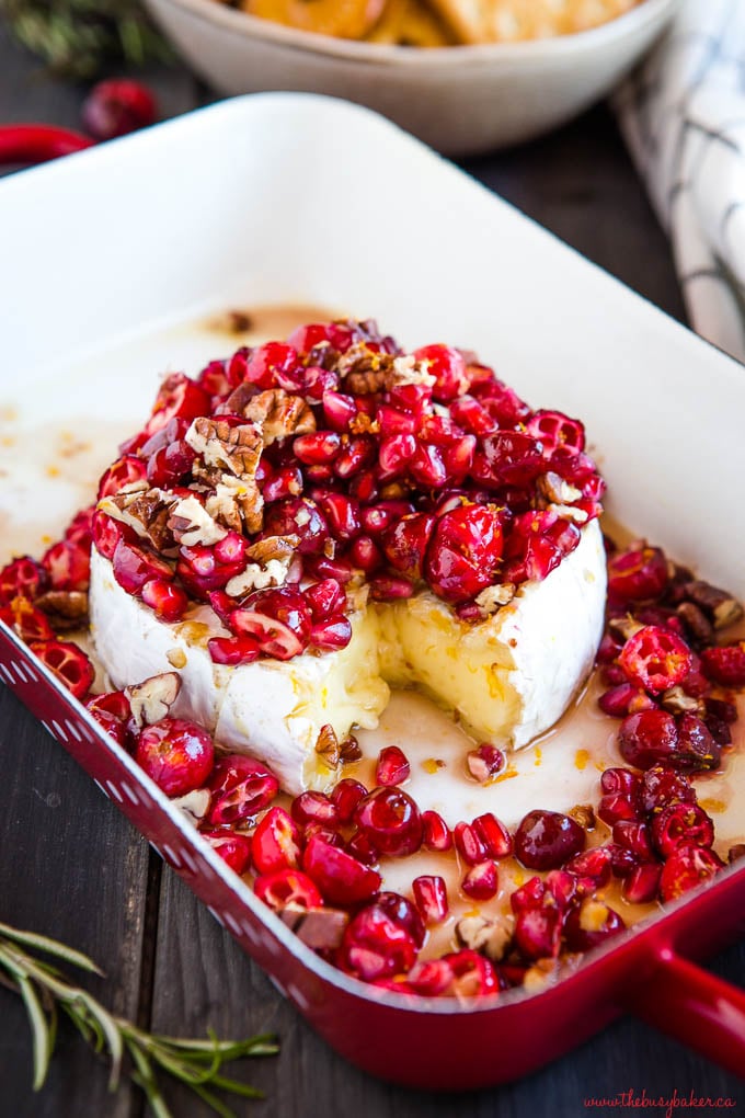 wheel of brie cheese topped with fresh cranberries and pomegranate arils in a red baking dish