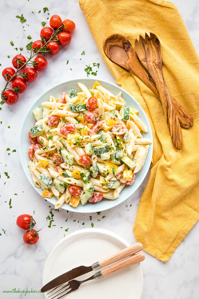 pasta salad in blue bowl with yellow kitchen towel, wood salad tongs and cherry tomatoes on the vine