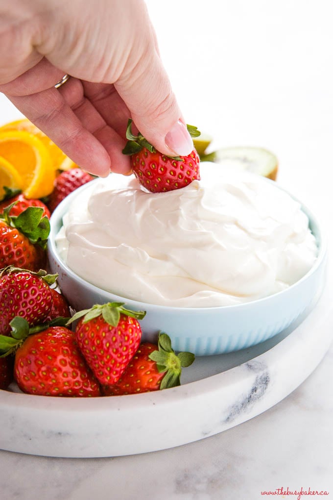 hand dipping strawberry into bowl of white fruit dip