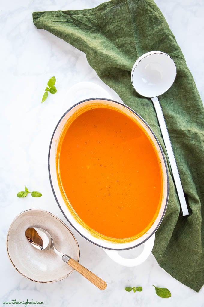 easy homemade creamy tomato soup in white pot with white ladle and basil leaves 
