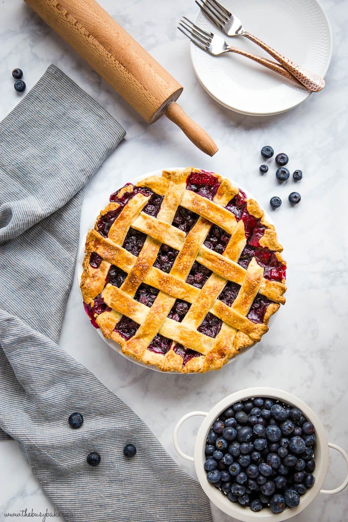 overhead image: blueberry pie with colander of blueberries, rolling pin, dessert plates with copper forks, and striped kitchen towel