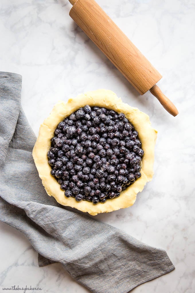 overhead image: raw pie crust filled with fresh blueberries, a rolling pin, and a striped kitchen towel