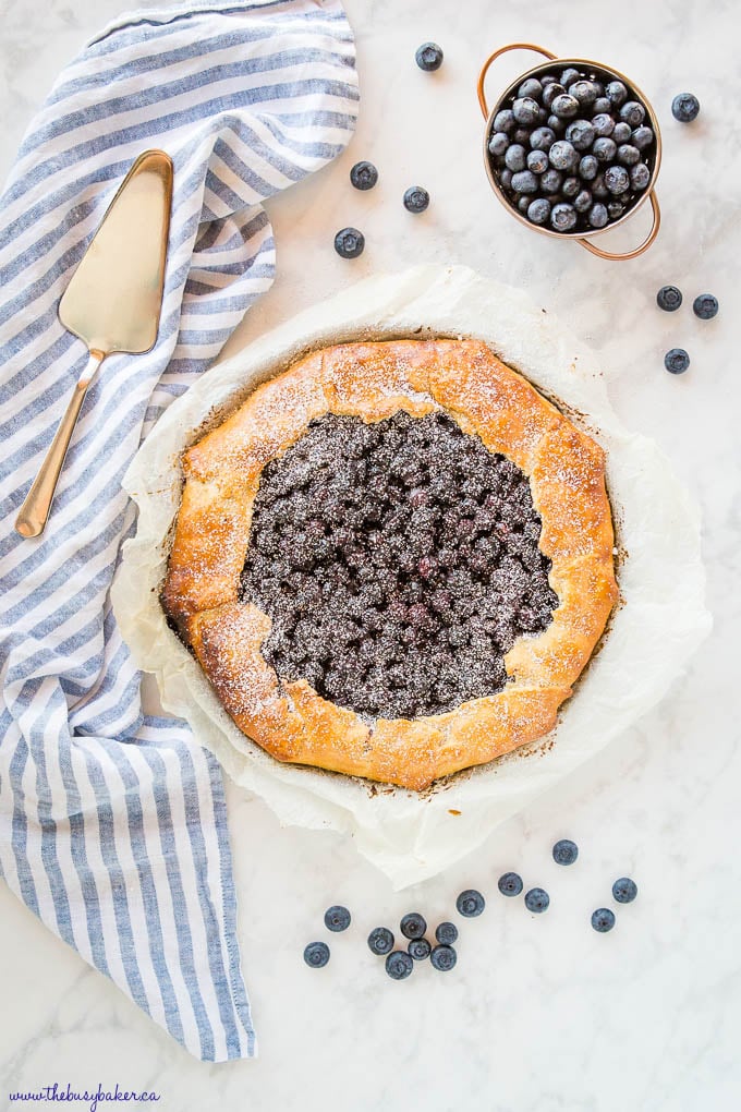overhead image: blueberry galette on parchment paper with blue striped kitchen towel and fresh blueberries
