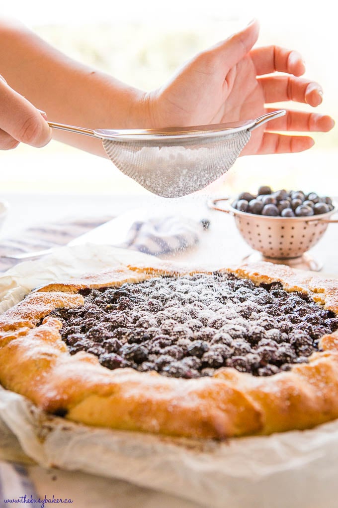 hand sprinkling powdered sugar over homemade blueberry galette with a small strainer