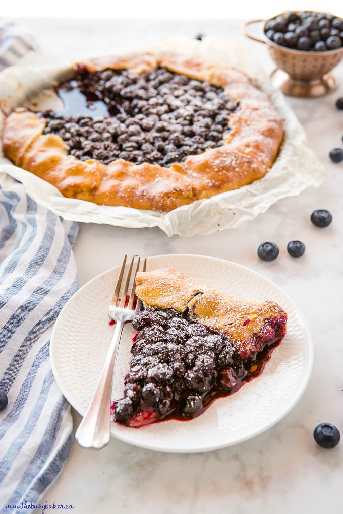 slice of blueberry galette on white plate with fork, with blueberry galette dessert in background, and fresh blueberries