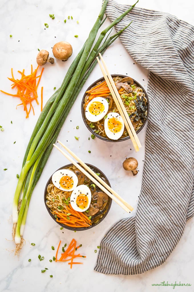 overhead image: 2 black ceramic bowls with ramen noodles, vegetables, and eggs, with green onions