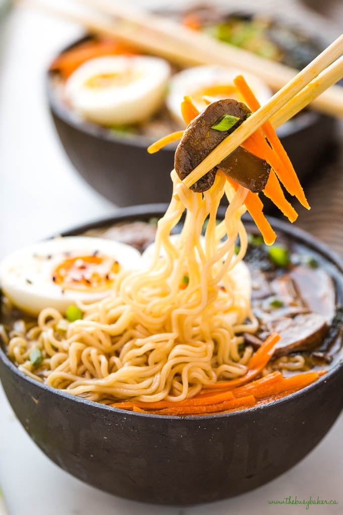 chopsticks lifting ramen noodles, mushroom and carrots out of black ceramic bowl