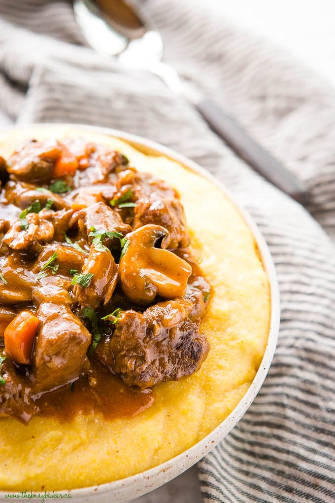 This Braised Beef and Mushrooms with Polenta is a comfort food dish that's easy to make and packed with savoury flavour! Creamy polenta with red wine braised tender beef and fresh mushrooms. Recipe from thebusybaker.ca! #comfortfood #beef #beefstew #braisedbeef #mushrooms #redwinebeef #polenta #homemade #recipe #familymeal close-up image: pottery bowl with polenta and braised beef and mushrooms