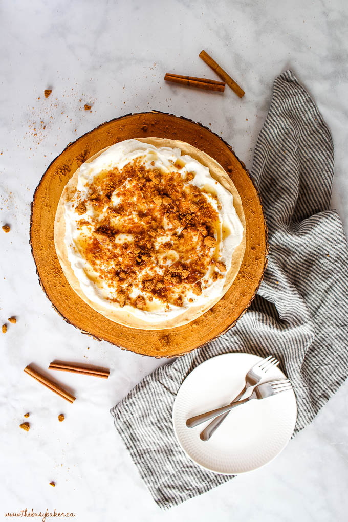 overhead image: no bake pumpkin cheesecake on wooden cake stand with whipped cream, cookie crumbs, and caramel sauce