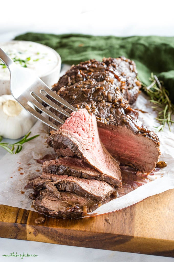 fork lifting a slice of beef tenderloin, pink on the inside with garlic, herbs and horseradish sauce in the background