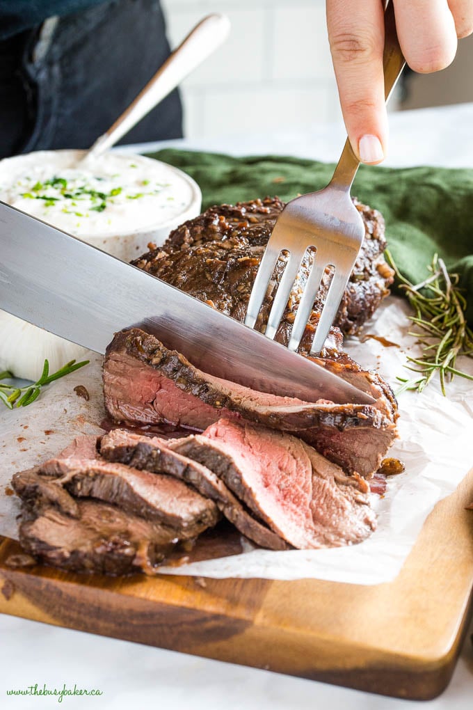 This Best Ever Marinated Beef Tenderloin is perfectly tender & juicy and packed with flavour - Easy to make with a simple marinade and great for the holidays served with horseradish sauce! Recipe from thebusybaker.ca! #holidays #beef #tenderloin #steak #horseradish #sauce #marinade #marinated #meat #roasted #christmas #holidays #newyears #celebration fork and knife slicing roasted beef tenderloin, medium rare