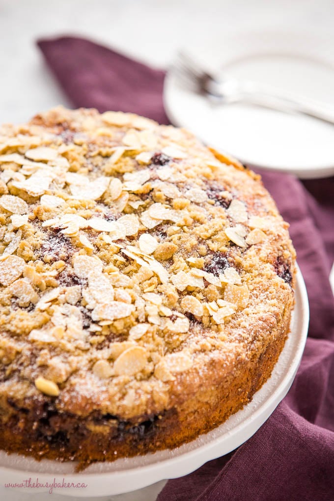 cherry almond coffee cake on white cake stand with slivered almonds and powdered sugar