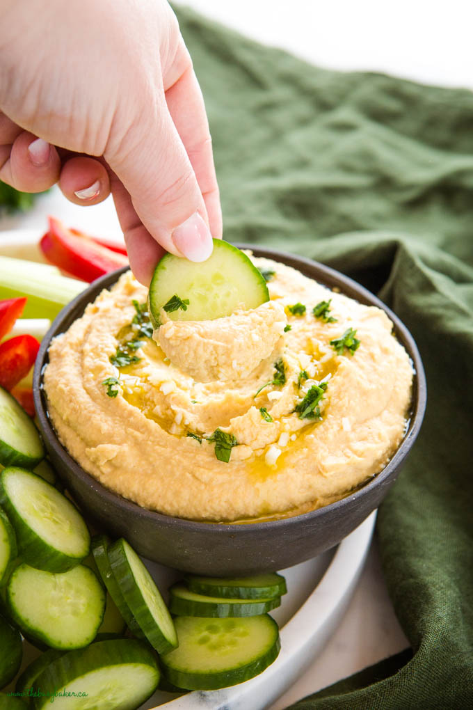 hand dipping cucumber slice in bowl of hummus