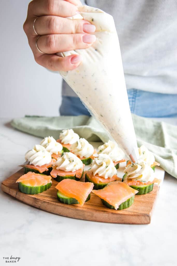 woman's hand piping cream cheese on cucumber slices and smoked salmon
