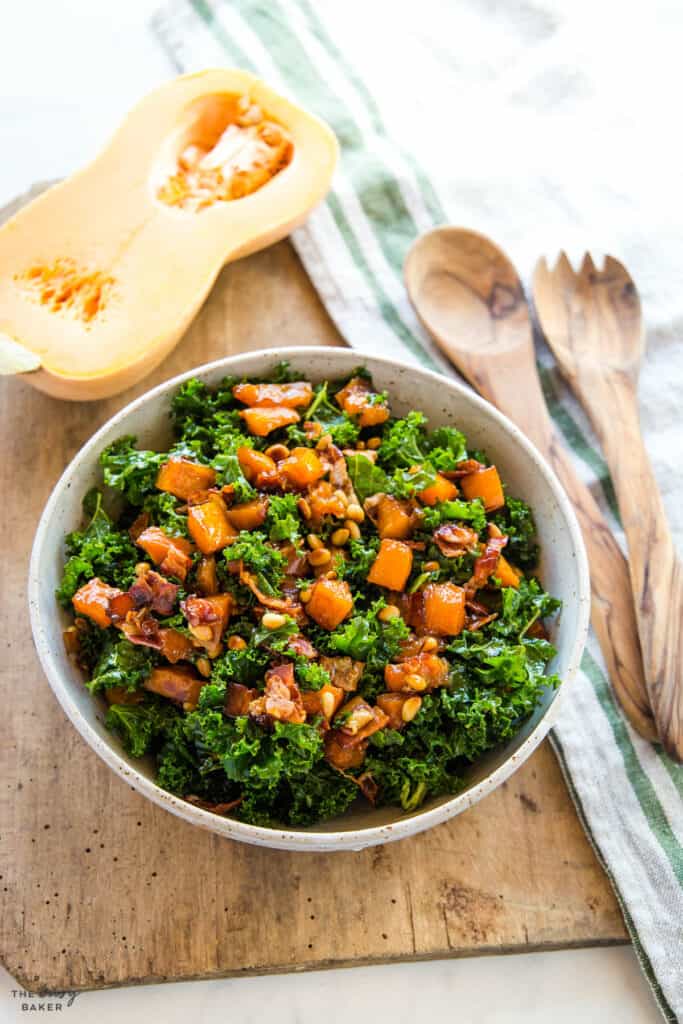  Kale, butternut Squash, pine nuts and bacon in a white bowl on a wood cutting board. half a butternut squash above.
