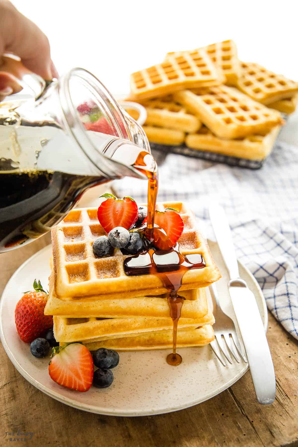 hand pouring syrup on buttermilk waffles in a stack with fruit 