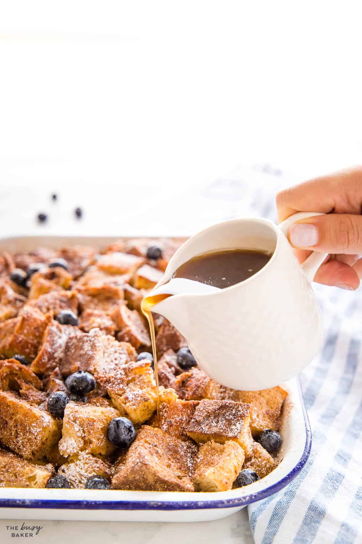hand pouring syrup on french toast casserole in a white baking pan with blue rim