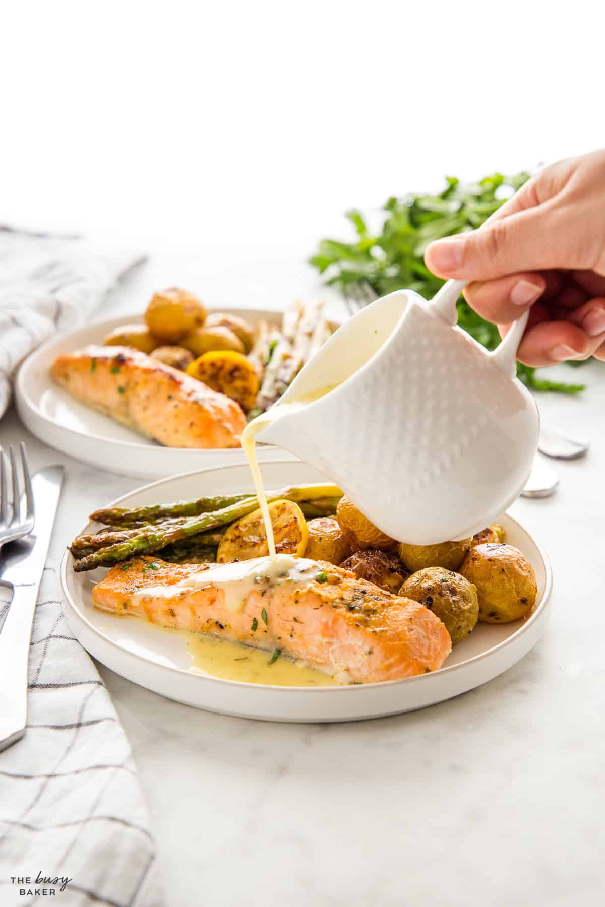 hand pouring lemon butter sauce over salmon and veggies on a white plate
