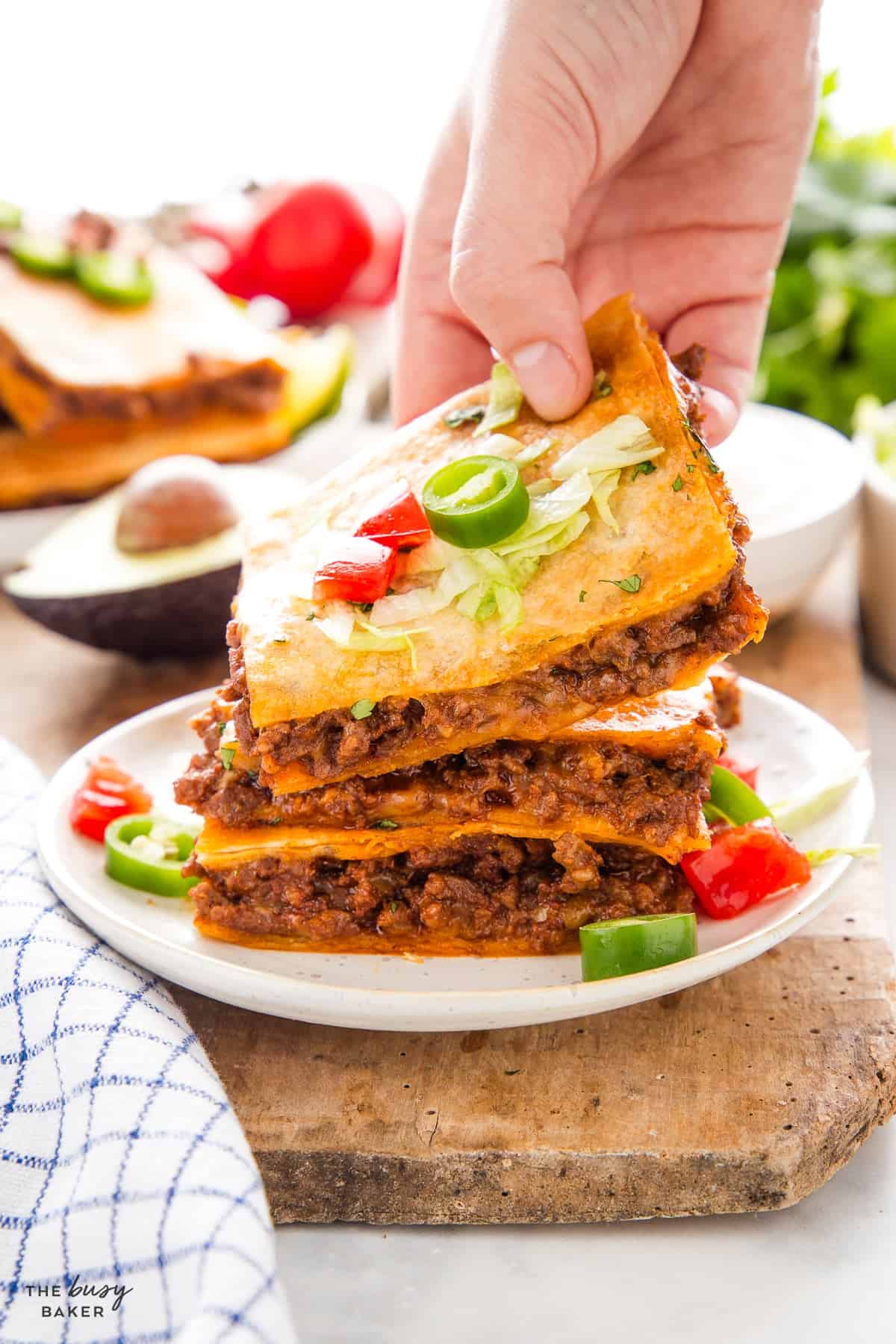 Hand reaching for a stack of sheet pan quesadilla squares showing layers of taco meat and cheese, topped with lettuce and jalapeño